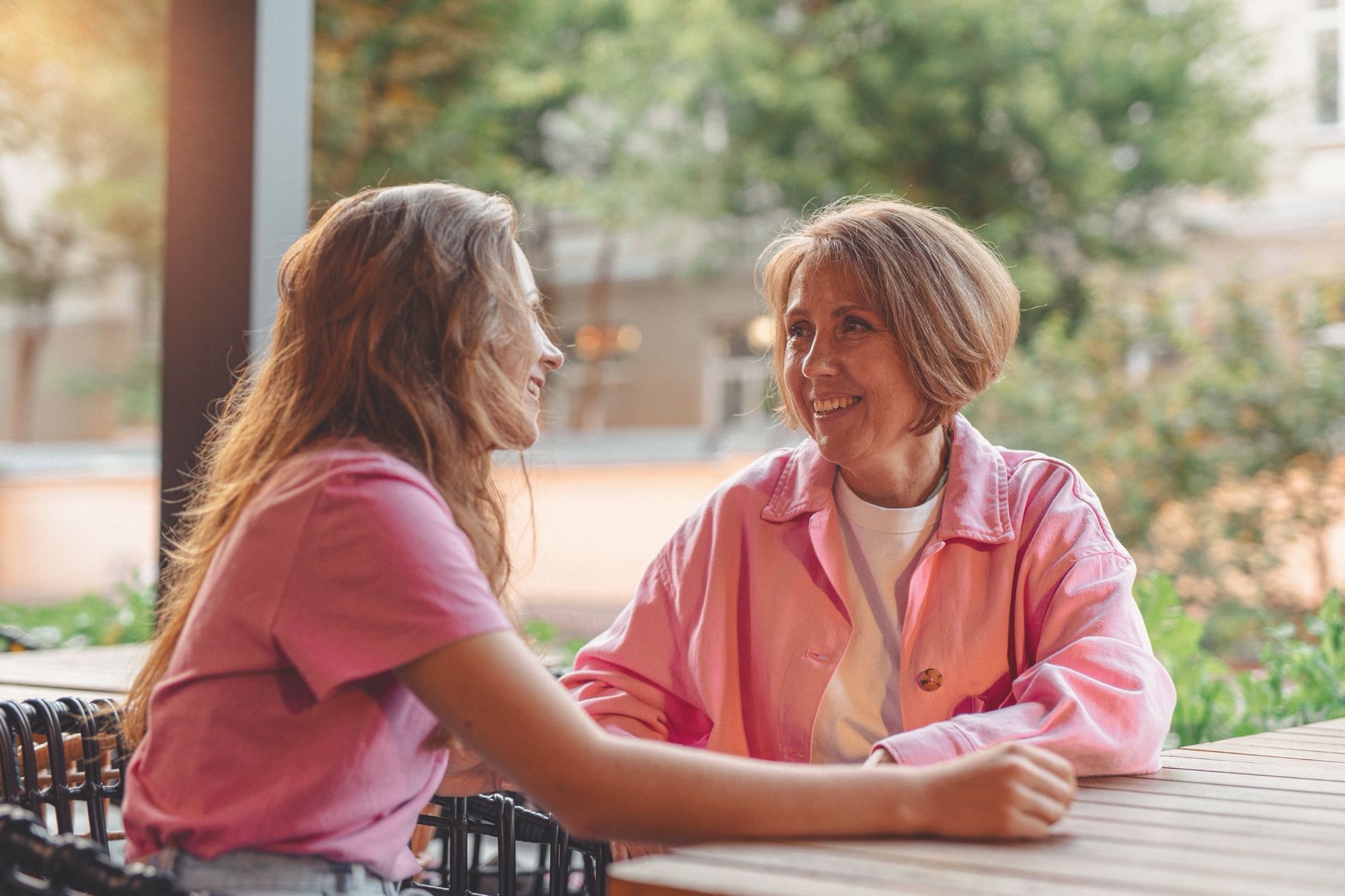 Two women at an outdoor table among green leaves, one talking, the other listening and smiling.