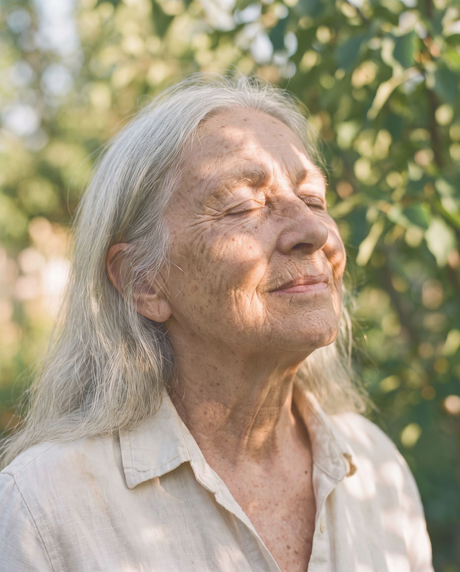 An older woman with long gray hair, eyes closed, smiling in the sun.