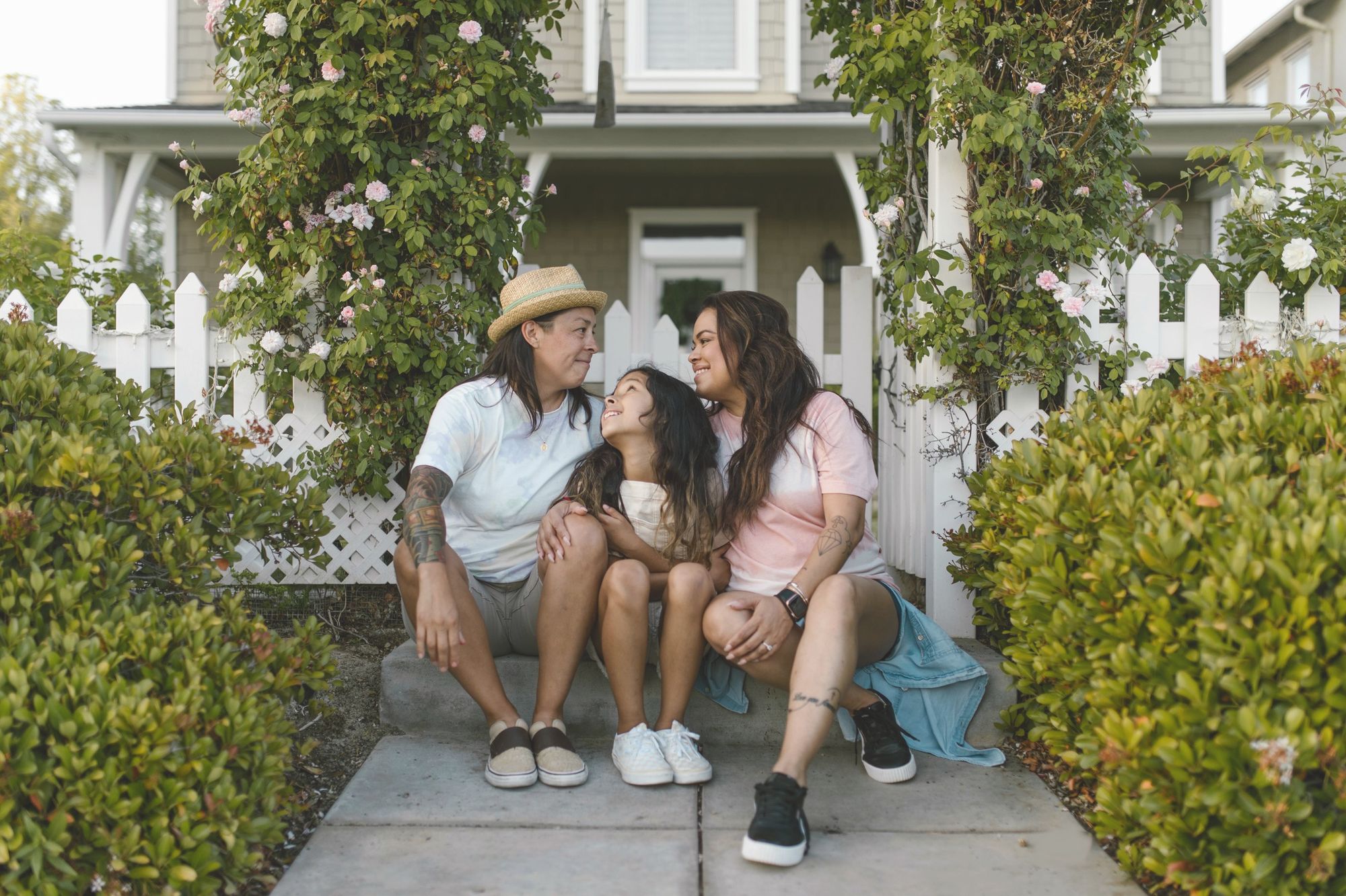 Two women and their daughter sitting close on a front walk among roses.
