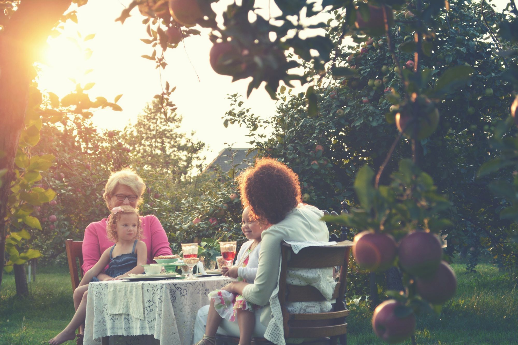 Two women and two small children at a table under an apple tree.